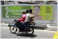 Three men on a motorcycle passing a glo billboard. Maroko Road, Victoria Island.